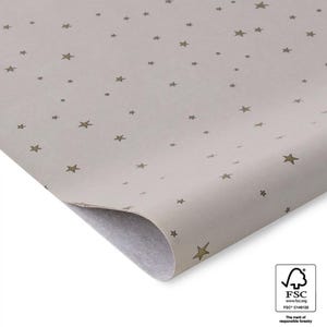 Könnte beinhalten: Ein Blatt hellbeiges Geschenkpapier mit einem sich wiederholenden Muster aus goldenen Sternen. Das Papier ist in der Mitte gefaltet und die Ecke ist nach oben gekrümmt. Das FSC-Logo ist in der unteren rechten Ecke sichtbar.