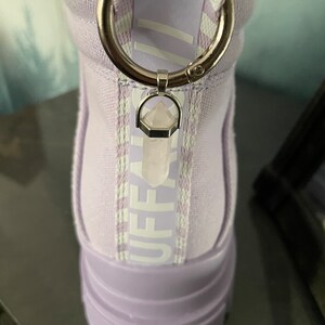 Könnte beinhalten: Lilafarbene High-Top-Sneaker mit einem weißen "Buffalo"-Logo an der Seite und einem weißen "BUFFALO"-Logo auf der Rückseite. Die Sneaker haben einen Kristall-Anhänger an der Reißverschlusslasche.