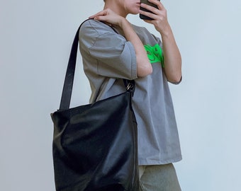May include: A person wearing a gray t-shirt with a green graphic, black sunglasses, and light-wash jeans is holding a black leather tote bag with a long strap over their shoulder.