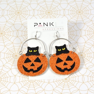 Puede incluir: Un par de pendientes de calabaza naranja con purpurina y caras de gato negro en la parte superior. Los pendientes están en una tarjeta blanca con el texto "P•NK XPRESSIONS".