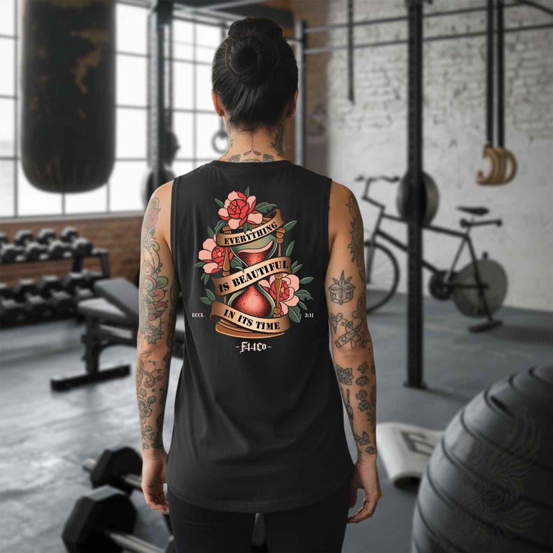 May include: Black tank top with a floral and hourglass design. The design includes the text "EVERYTHING IS BEAUTIFUL IN ITS TIME" on a banner, with the brand name "FLCO" below. The tank top is sleeveless.
