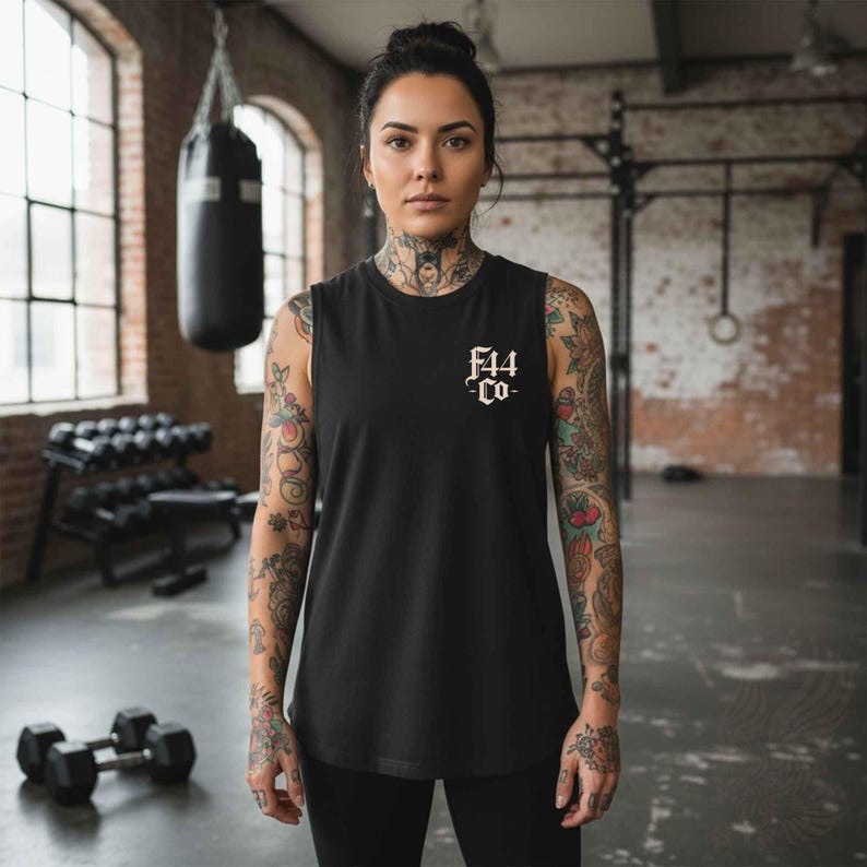 May include: Black sleeveless tank top with the text "F44 Co-" in white script. The tank top is worn by a person in a gym setting. The person has tattoos on their arms and neck. Dumbbells and a punching bag are visible in the background.