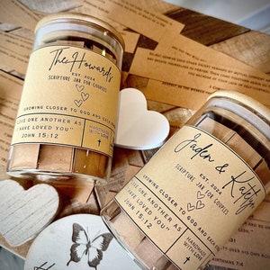 May include: Two glass jars with wooden lids filled with small pieces of paper. The jars are labeled "The Howards Scripture Jar for Couples" and "Jaden & Katlyn Scripture Jar for Couples". Both jars have a brown label with white text and a small cross symbol.