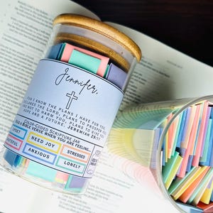May include: A glass jar with a wooden lid filled with colorful paper slips. The jar is labeled "Jennifer." and has a cross symbol. The jar is on a book with the text "FOR I KNOW THE PLANS I HAVE FOR YOU, DECLARES THE LORD, PLANS TO PROSPER YOU AND NOT TO HARM YOU, PLANS TO GIVE YOU HOPE AND A FUTURE. JEREMIAH 29:11" printed on it. The jar is a color-coded scripture jar with a label that reads "Color-Coded Scripture Jar Read a Bible Verse When You Are Feeling..." and has color-coded squares for "Upset", "Grateful", "Need", "Joy", "Anxious", "Stressed", and "Lonely".