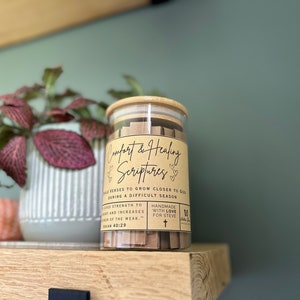 May include: A glass jar filled with small brown paper strips containing bible verses. The jar has a brown label that reads "Comfort & Healing Scriptures" with the text "Bible verses to grow closer to God during a difficult season".