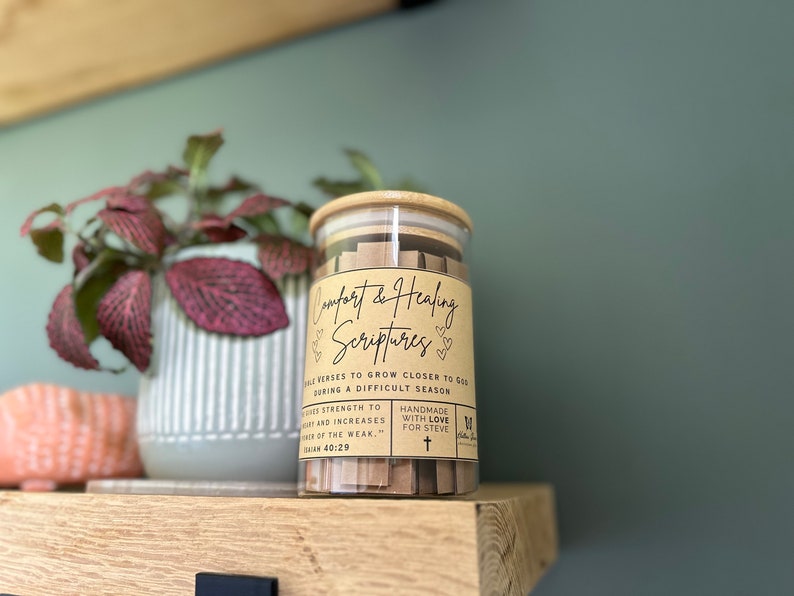 May include: A glass jar filled with small brown paper strips containing bible verses. The jar has a brown label that reads "Comfort & Healing Scriptures" with the text "Bible verses to grow closer to God during a difficult season".