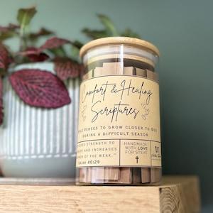 May include: A glass jar with a wooden lid filled with small pieces of paper. The jar has a label that says "Comfort & Healing Scriptures" and includes the text "Bible verses to grow closer to God during a difficult season." The label also says "Handmade with love for Steve".