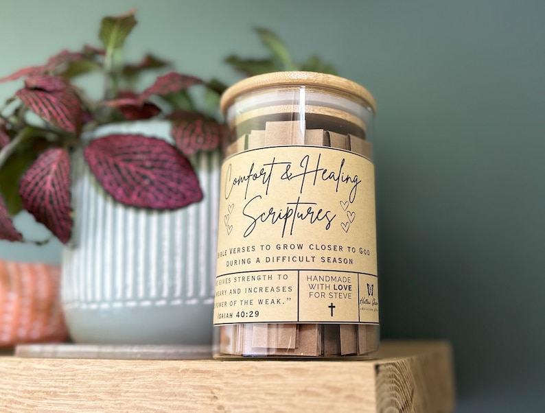 May include: A glass jar with a wooden lid filled with small pieces of paper. The jar has a label that says "Comfort & Healing Scriptures" and includes the text "Bible verses to grow closer to God during a difficult season." The label also says "Handmade with love for Steve".