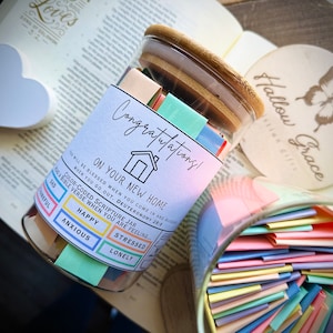 May include: A glass jar filled with colorful slips of paper, labeled "Color-Coded Scripture Jar". The jar is decorated with a house icon and the text "Congratulations! On Your New Home". The jar is sitting on a book with the text "Your Soul Loves" visible.