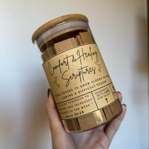 May include: A glass jar filled with small pieces of paper with bible verses printed on them. The jar has a brown paper label that reads "Comfort & Healing Scriptures" with hearts and a cross. The label also includes the text "Bible Verses to Grow Closer to God During a Difficult Season" and "Handmade with Love for Nancy".