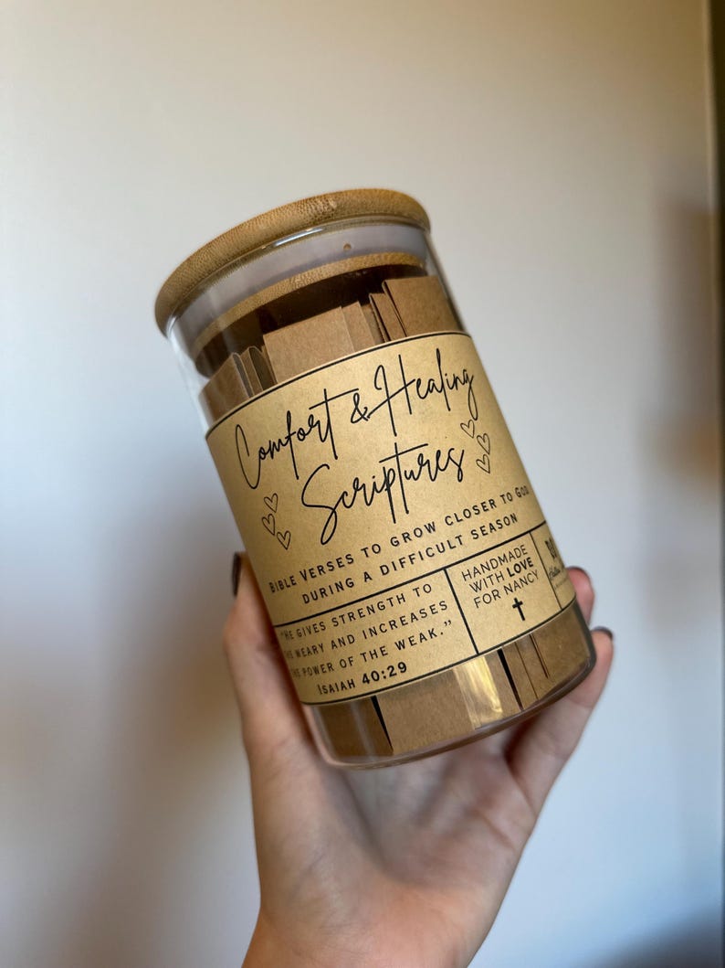 May include: A glass jar filled with small pieces of paper with bible verses printed on them. The jar has a brown paper label that reads "Comfort & Healing Scriptures" with hearts and a cross. The label also includes the text "Bible Verses to Grow Closer to God During a Difficult Season" and "Handmade with Love for Nancy".