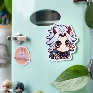 May include: A sticker of a cartoon character with white hair, red eyes, and horns. The character is wearing a black and purple outfit and is holding out their hand. The sticker is on a light blue refrigerator.
