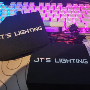 Puede incluir: Dos cajas rectangulares negras con el texto "JT's Lighting" en letras blancas. Las cajas est&aacute;n conectadas por un haz de cables con conectores.