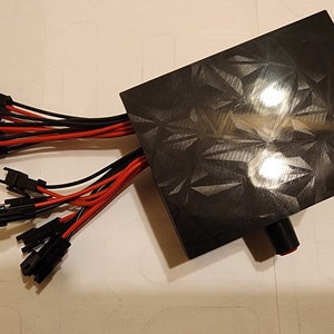 Puede incluir: Una caja de pl&aacute;stico negra con una superficie texturizada y un bot&oacute;n rojo. M&uacute;ltiples cables negros y rojos con conectores se extienden desde la caja.