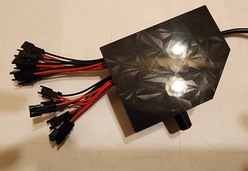 Puede incluir: Una caja de pl&aacute;stico negra con una superficie texturizada y un bot&oacute;n rojo. M&uacute;ltiples cables negros y rojos con conectores se extienden desde la caja.