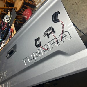 Puede incluir: Un port&oacute;n trasero de cami&oacute;n plateado con la palabra "TUNDRA" en relieve en el metal. Tres accesorios de luz de pl&aacute;stico negro est&aacute;n unidos al port&oacute;n trasero con cables rojos y blancos.