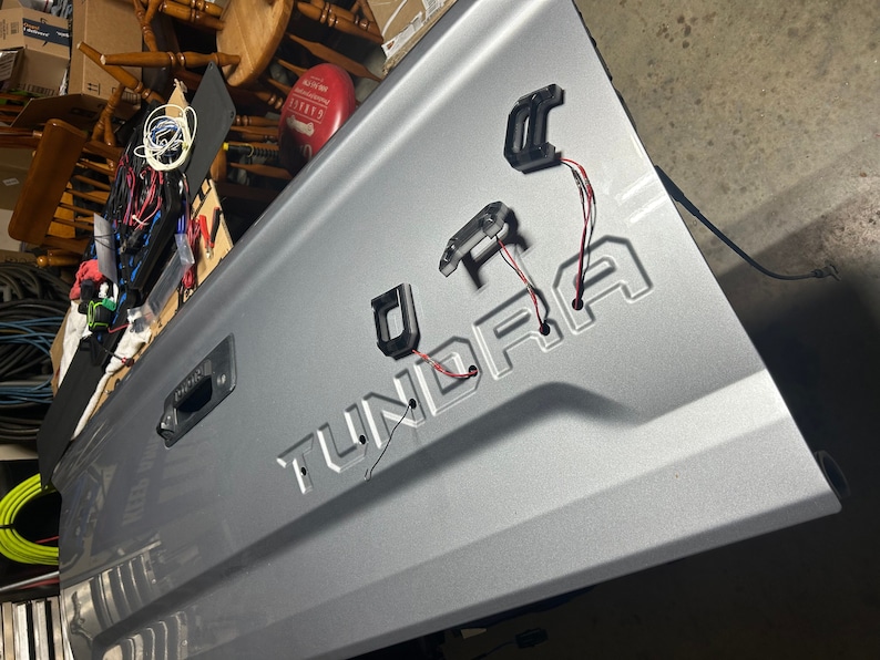 Puede incluir: Un port&oacute;n trasero de cami&oacute;n plateado con la palabra "TUNDRA" en relieve en el metal. Tres accesorios de luz de pl&aacute;stico negro est&aacute;n unidos al port&oacute;n trasero con cables rojos y blancos.