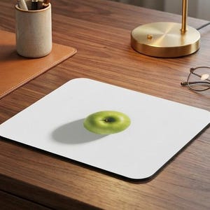 May include: A white square mousepad with a green apple in the center. The mousepad sits on a wooden desk, alongside a brass desk lamp, a succulent plant, and a pair of glasses. A leather notebook and pen holder are also visible.