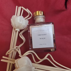 May include: A clear glass diffuser bottle with a gold cap, labeled "ORCHARD WALK" and 6 fl oz / 177 mL.  It is surrounded by decorative reed sticks and white flower-shaped diffusers, all set against a red background.