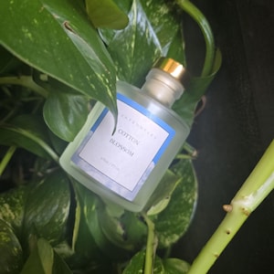 May include: A clear glass perfume bottle with a gold cap and a label that reads "Cotton Blossom" is nestled among green leaves. The bottle contains 6 fl oz / 177 ml of product. The background is dark, highlighting the bottle and foliage.