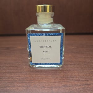 May include: A clear, square glass bottle with a gold cap and a label that reads "SCENTEDBYLEY TROPICAL VIBE 6 fl oz / 177 mL." The label has a blue and white design. The bottle is on a brown surface.
