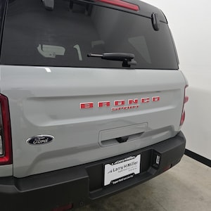 Puede incluir: Vista trasera de un SUV Ford Bronco Sport gris claro con parachoques trasero negro, ventana trasera tintada con limpiaparabrisas, letras rojas Bronco Sport en el port&oacute;n trasero, emblema Ford y marco de matr&iacute;cula de Larry H Miller.