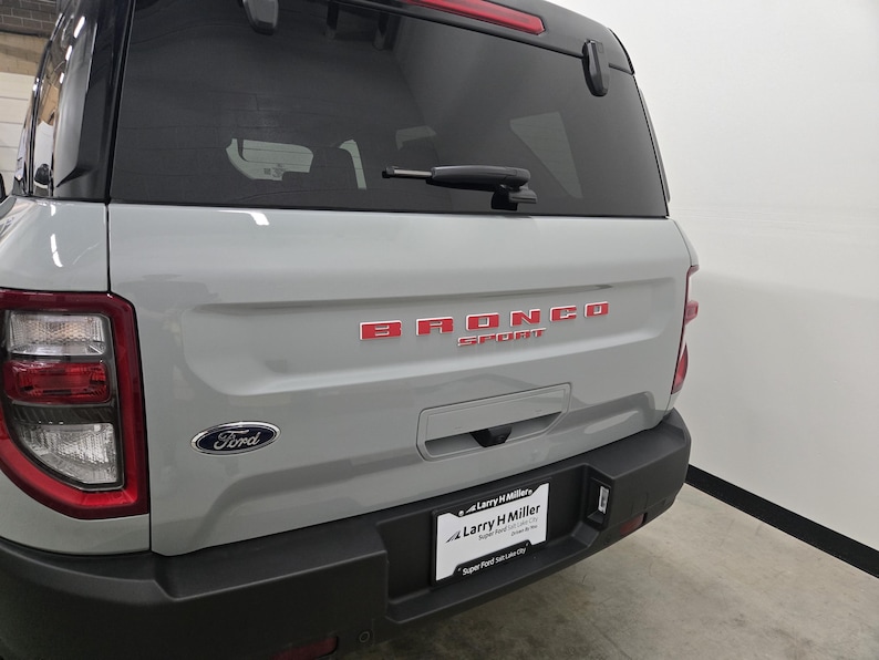 Puede incluir: Vista trasera de un SUV Ford Bronco Sport gris claro con parachoques trasero negro, ventana trasera tintada con limpiaparabrisas, letras rojas Bronco Sport en el port&oacute;n trasero, emblema Ford y marco de matr&iacute;cula de Larry H Miller.