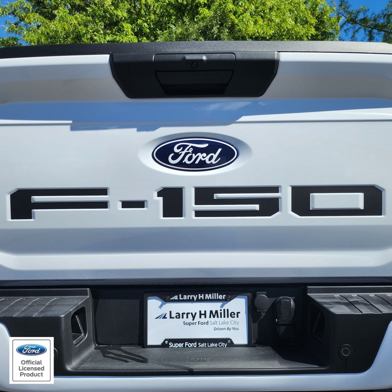Op de afbeelding: Witte Ford F-150-truckachterklep met zwarte letters en het Ford-logo. De kentekenplaathouder luidt "Larry H Miller Super Ford Salt Lake City". Een officieel gelicentieerd productbadge staat linksonder.