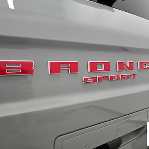 May include: A light gray vehicle tailgate featuring the raised lettering BRONCO in bold red with white outlines, positioned above the word SPORT in a smaller red font. A small white rectangular logo in the corner reads Ford Official Licensed Product.