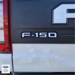Puede incluir: Un portón trasero de una camioneta Ford F-150 blanca con un panel de plástico negro que muestra el logotipo de Ford y el texto "F-150".
