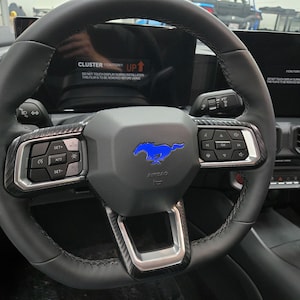 Può includere: Un volante in pelle nera con un logo Mustang blu e dettagli in fibra di carbonio. Il volante ha controlli per il sistema audio dell'auto e il cruise control.