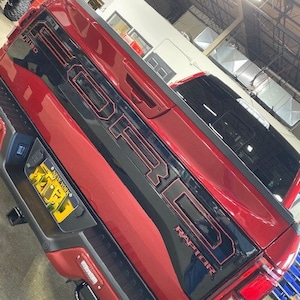 Peut inclure: Un camion Ford Raptor rouge avec un hayon noir et le mot "RAPTOR" en lettres rouges. Le camion a une plaque d'immatriculation de New York.