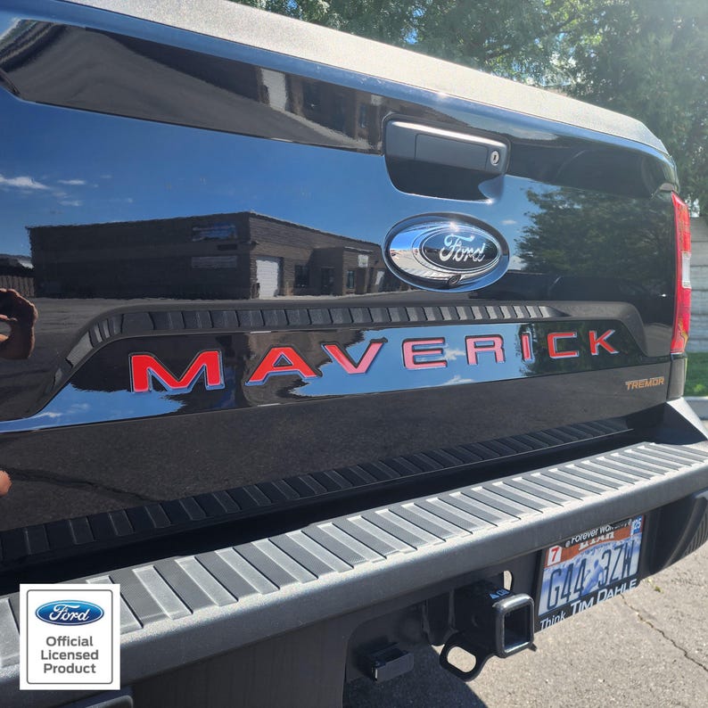 May include: Black Ford Maverick truck tailgate with red "MAVERICK" lettering and a chrome Ford emblem. The truck has a black bumper and a license plate visible. The image is an official licensed Ford product.
