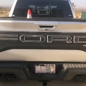 Peut inclure: Un camion Ford Raptor argent&eacute; avec un hayon noir et le mot "RAPTOR" en lettres noires sur le hayon.