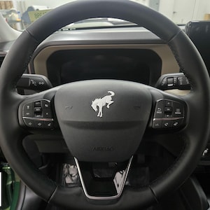 May include: Black leather steering wheel with integrated controls. The center features a silver emblem of a horse. The word "AIRBAG" is visible. The Ford logo is in the lower left corner.