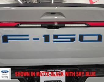 May include: A close-up of the back of a gray Ford F-150 pickup truck. The tailgate has a black "F-150" emblem with a blue outline. The text "SHOWN IN MATTE BLACK WITH SKY BLUE" is visible below the emblem.