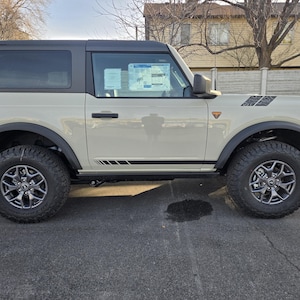 May include: A light tan SUV with a black roof and black detailing. The vehicle has large, rugged tyres and a spare tyre mounted on the back. The vehicle is parked on a paved surface.