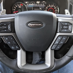 May include: A black leather steering wheel with silver accents and controls. The steering wheel has a silver "RAPTOR" emblem in the center.
