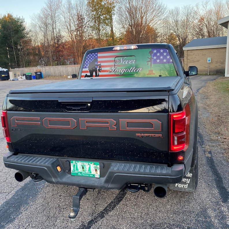 Peut inclure: Un pick-up Ford Raptor noir avec un autocollant sur la lunette arri&egrave;re qui dit "Never Forgotten" et repr&eacute;sente un drapeau am&eacute;ricain et la silhouette d'un soldat. Le camion a un logo Ford rouge et une plaque d'immatriculation.