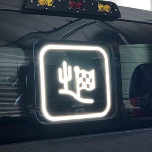 May include: White decal sticker with a cactus and chequered flag design on a black background. The sticker is in a square frame with a white border.