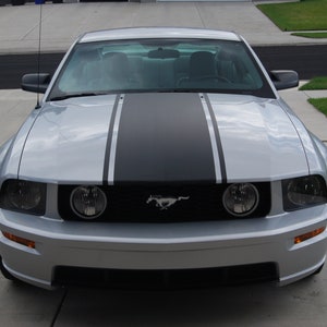 Puede incluir: Un Ford Mustang plateado con rayas de carreras negras en el capó. El coche tiene una parrilla delantera negra con un logotipo de Mustang cromado.
