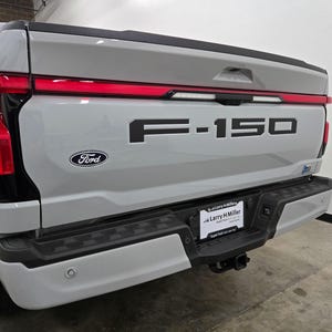 Puede incluir: Una camioneta Ford F-150 gris con una puerta trasera negra y un emblema "F-150" negro. La camioneta tiene una luz trasera roja y un parachoques negro.