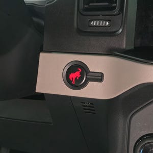 Może przedstawiać: Zbliżenie wnętrza samochodu z czarną deską rozdzielczą i beżowym akcentem. Podświetlane na czerwono logo Bronco znajduje się na czarnym przycisku. Logo Ford Official Licensed Product znajduje się w lewym dolnym rogu.