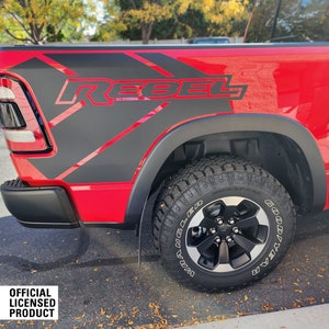 Op de afbeelding: Een rode pick-up truck met zwarte spatborden en een zwarte "Rebel" sticker aan de zijkant van de laadbak.