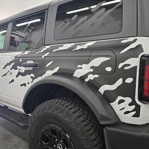 May include: A white SUV with a black and white camouflage pattern wrap. The vehicle has blacked-out windows and a black tire with a black rim.