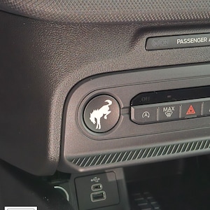 May include: A detailed shot of a car's interior, focusing on a black button with a white horse emblem. The dashboard includes various function buttons and USB ports. The Ford logo is displayed in the corner of the image.