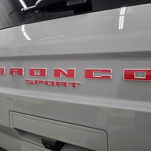 Puede incluir: Un port&oacute;n trasero de veh&iacute;culo de color gris claro con la palabra BRONCO en letras rojas en relieve con un borde blanco. Debajo de la palabra BRONCO, la palabra SPORT aparece en letras rojas m&aacute;s peque&ntilde;as y a juego con borde blanco.