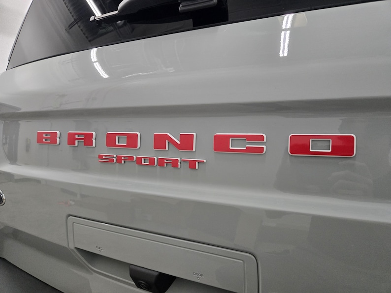 Puede incluir: Un port&oacute;n trasero de veh&iacute;culo de color gris claro con la palabra BRONCO en letras rojas en relieve con un borde blanco. Debajo de la palabra BRONCO, la palabra SPORT aparece en letras rojas m&aacute;s peque&ntilde;as y a juego con borde blanco.