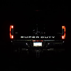May include: Rear view of a black pickup truck with the words "SUPER DUTY" in white lettering on the tailgate. The taillights are red and the truck is parked in a dark setting.