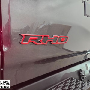 May include: A close-up of a black truck tailgate with a red "RHO" emblem. The emblem is outlined in black and has a raised, three-dimensional appearance. The truck is a dark gray color and has a black bumper.